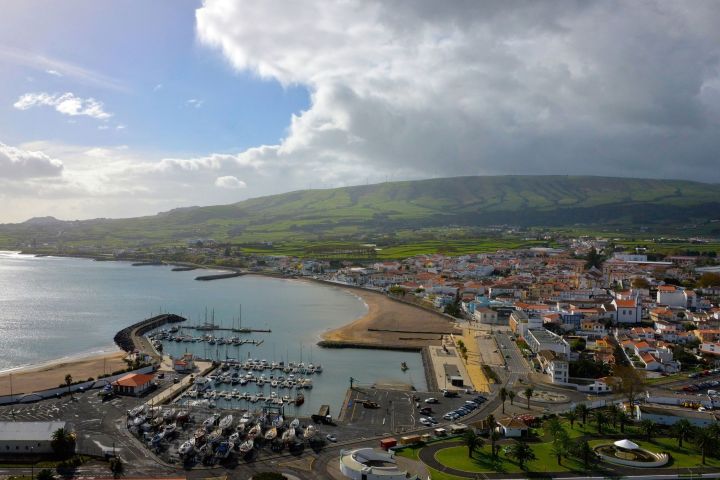 Aerial view of a coastal town with a marina, boats, and green hills under a cloudy sky.