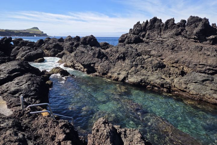 Rocky coastal pool with clear water and metal railings under a blue sky.