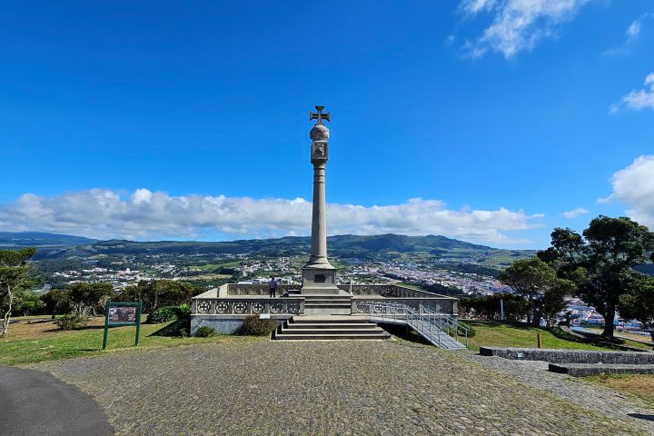 Tall pillar with cross on hilltop, overlooking town in valley under blue sky.