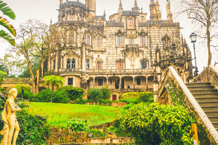 Ornate mansion with spires, surrounded by lush garden and stone stairs.