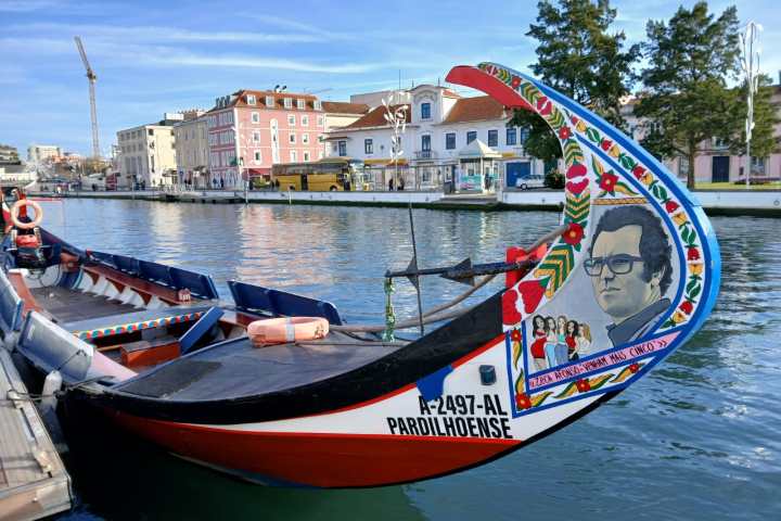 Colorful boat docked by canal with painted portrait and flowers, cityscape in background.