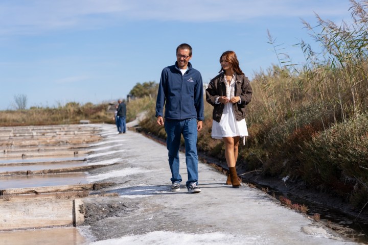 Two people walking on a path by salt pans, surrounded by grass, under a clear blue sky.