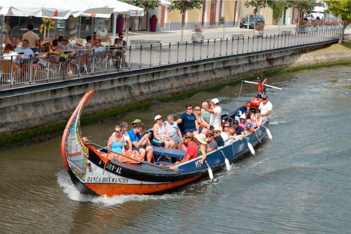 Tourists in a colorful gondola-like boat on a canal with people seated at a riverside cafe.