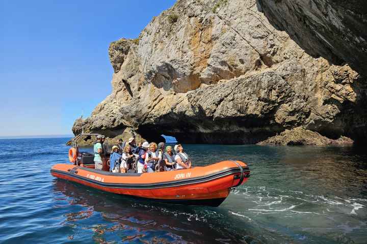 People in red boat near large rocky cliff on clear, sunny day.