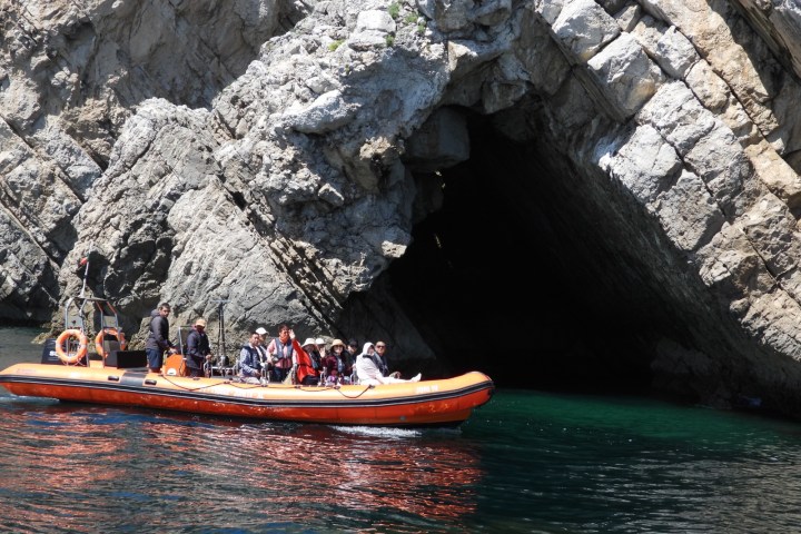 Boat with people near cave entrance in rocky cliff over water.