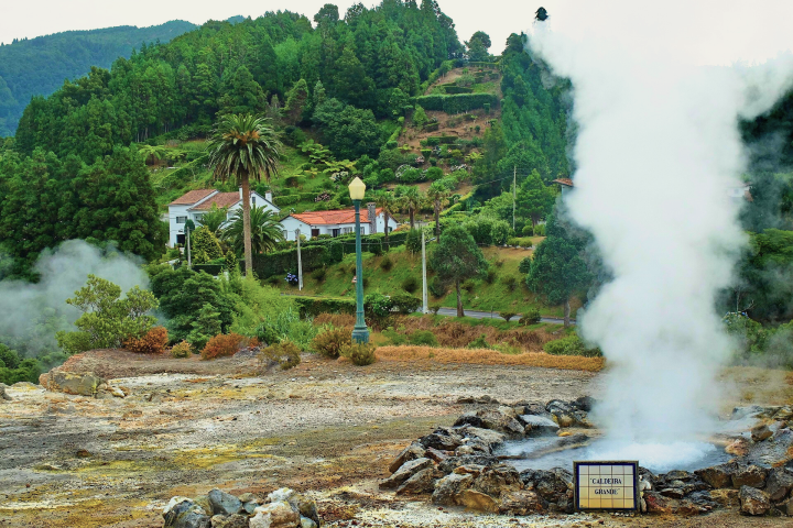 Steam rises from a geyser in a lush, hilly landscape with houses.