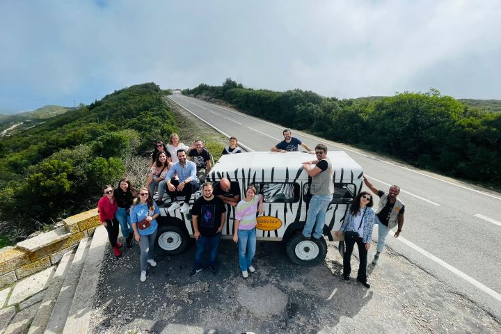 Group of people posing on and around a zebra-striped vehicle on a road with greenery and a blue sky.