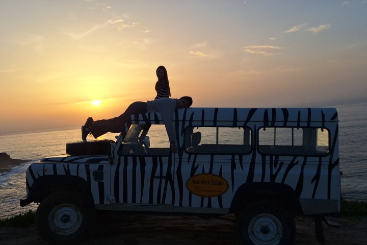 Silhouetted people on zebra-striped vehicle at sunset by ocean.