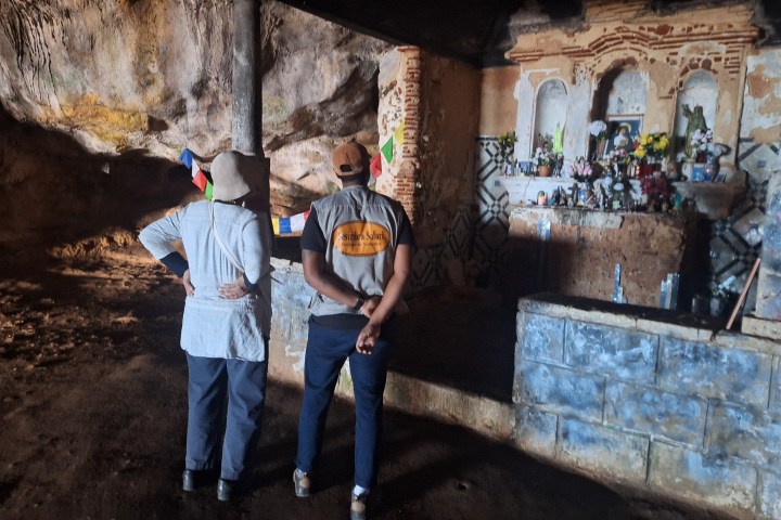 Two people in a cave, observing a small shrine with colorful flags.