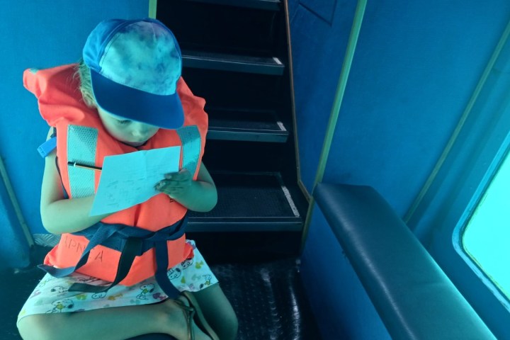 Child in a life jacket writing on paper inside a blue boat cabin.