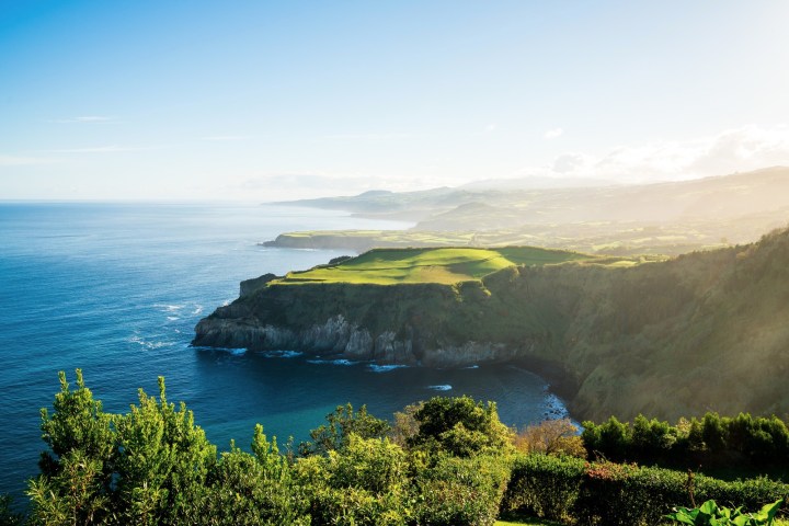 Coastal cliffs with green fields and ocean under a clear blue sky.