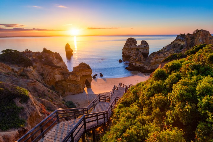 Sunset over a rocky beach with a wooden path and lush vegetation.