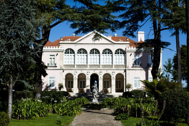 Elegant pink mansion with arched windows, surrounded by trees and gardens on a sunny day.
